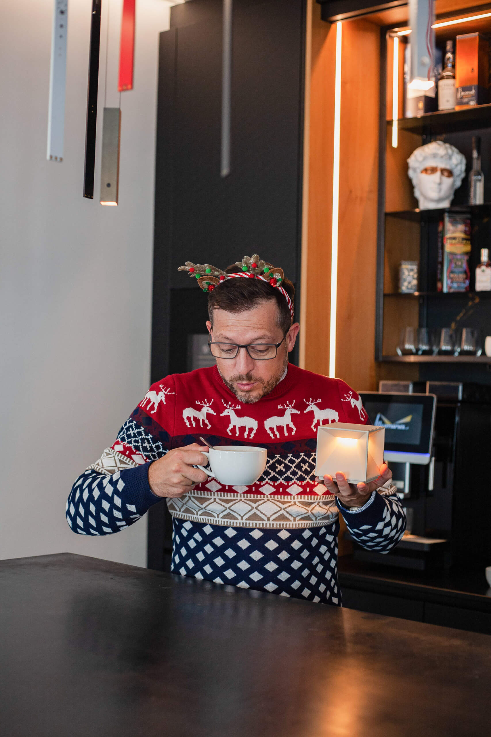 Mann im Weihnachtspullover mit Rentier-Haarreif hält Kaffee und Kerze