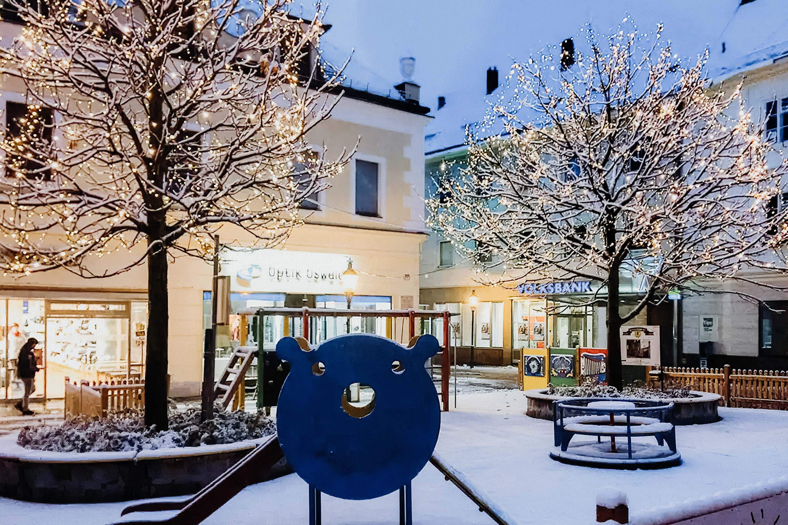 Schneebedeckter Spielplatz mit Lichterketten, Geschäften und Bank im Hintergrund.